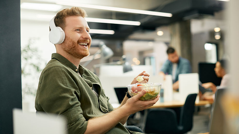 Man enjoying eating lunch, wearing headphones in the office.